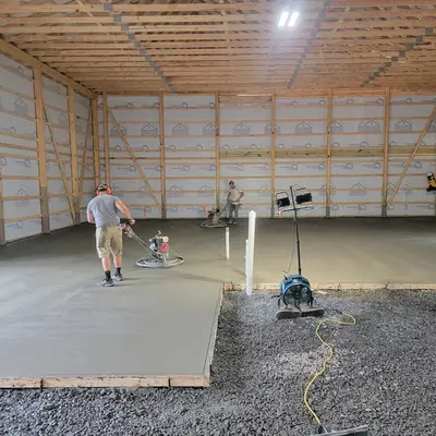 A worker power-troweling a freshly poured slab inside a pole building.